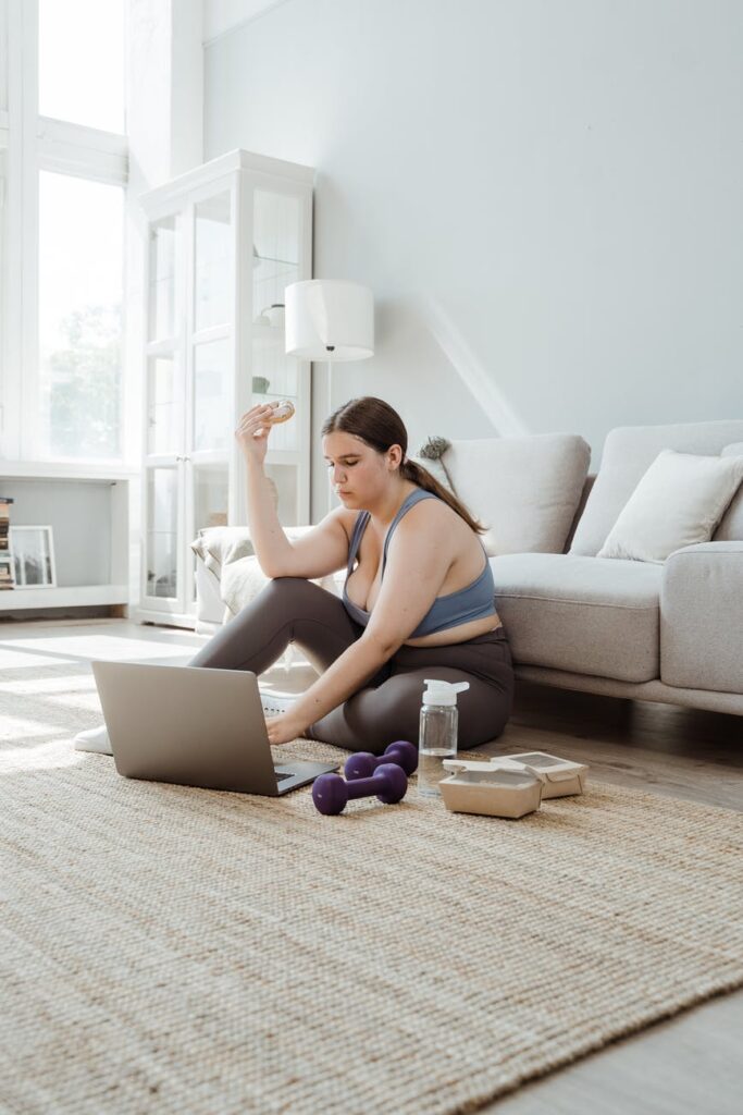 pcos weight loss - woman in sportswear sitting on floor using laptop and eating doughnut